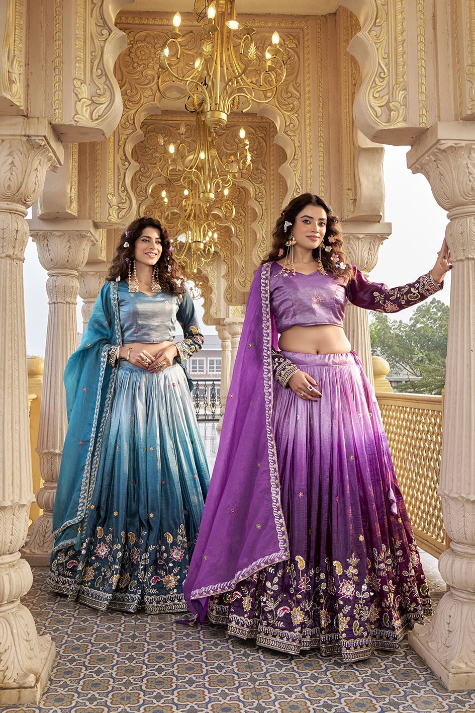 Two women in traditional blue and purple lehengas standing in an ornate indoor setting.